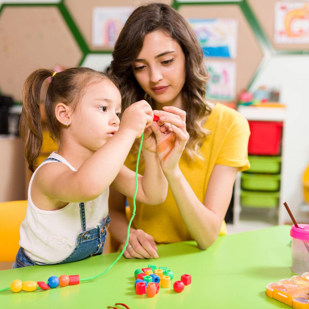 preschool teacher helping student string beads
